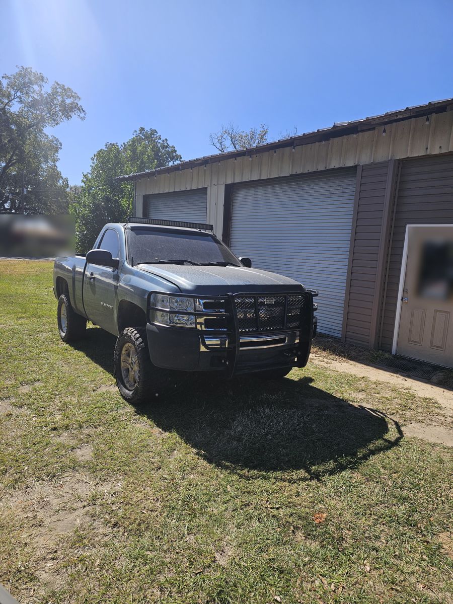 Blue-gray 2008 Chevy Silverado with a grille guard, parked on grass by a metal shop building